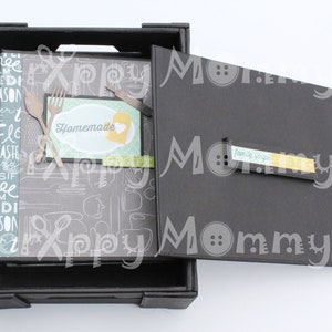 May include: A black wooden box with a lid that opens to reveal a recipe book with a gray and white pattern. The book has a label that says "Homemade" and a heart. The lid has a yellow label that says "Family Recipes".