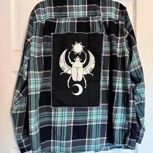 Puede incluir: Camisa de franela a cuadros en verde azulado, negro y gris. Un parche negro en la espalda presenta un diseño de escarabajo blanco con un sol y una luna creciente. La camisa está colgada en una puerta blanca.