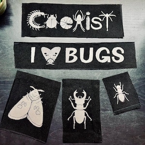 Puede incluir: Parches de tela negra con ilustraciones de insectos blancos y texto. Un parche dice "COEXIST" con dibujos de insectos. Otro dice "I BUGS" con un insecto en forma de corazón. Otros parches presentan una polilla, un escarabajo y una hormiga.