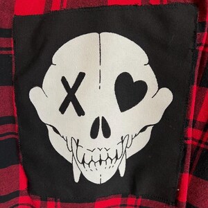 Puede incluir: Camisa de franela a cuadros rojos y negros con un parche negro. El parche muestra un diseño de calavera blanca, con una X para un ojo y un corazón para el otro. La camisa es de estilo casual.