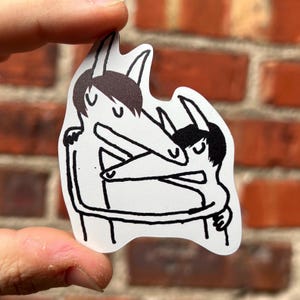 May include: A white sticker featuring a black line drawing of two figures embracing. The figures have long noses, black hair, and are hugging each other. The sticker is held up against a red brick wall.