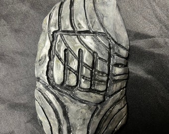 Pode incluir: Um objeto retangular cinza esculpido com um design de punho. O objeto tem uma superfície texturizada com contornos cinza escuro e parece ser feito de pedra ou material semelhante. O punho está no centro do objeto.