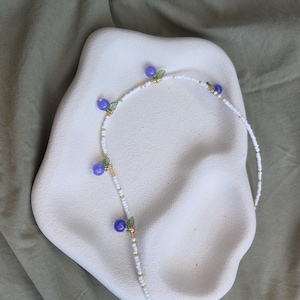 May include: A beaded necklace featuring a strand of small white seed beads interspersed with gold-coloured spacers. The necklace is accented with four purple spherical beads, each paired with a small green leaf-shaped charm attached by gold wire.