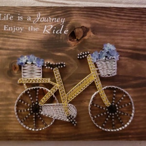 May include: A yellow bicycle with a basket of blue flowers on the handlebars. The bicycle is made of string wrapped around nails on a wooden board. The text "Life is a Journey Enjoy the Ride" is written above the bicycle.