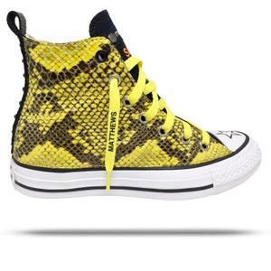 May include: High-top sneaker with a yellow and brown snakeskin pattern. The shoe has yellow laces with the name "MATTHEWS" printed on them. The sole is white with a black stripe, and the toe cap features a white star.