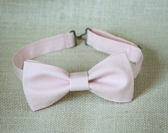 May include: A light pink bow tie made of fabric. The bow tie is tied and ready to wear.