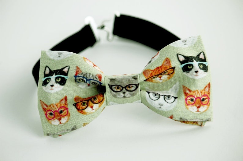 May include: A green bow tie with a pattern of cats wearing glasses. The bow tie is made of fabric and has an elastic band.