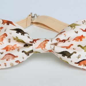 以下が含まれることがあります： 茶色、緑、オレンジ色の恐竜の繰り返し模様が特徴のクリーム色の蝶ネクタイ。ベージュの調節可能なネックストラップが付いています。デザインは気まぐれで、子供に適しています。蝶ネクタイは布製です。