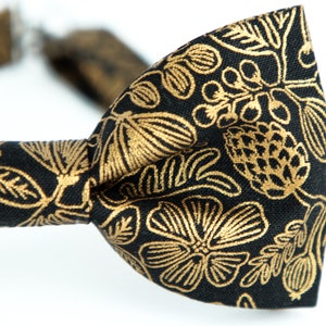 Puede incluir: Una pajarita negra con un patr&oacute;n floral dorado. El patr&oacute;n presenta hojas, flores y bayas.