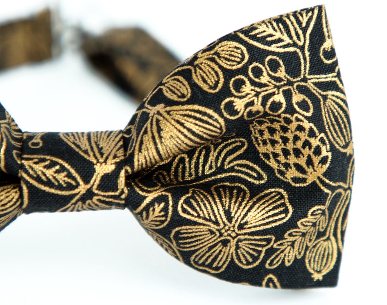 Puede incluir: Una pajarita negra con un patr&oacute;n floral dorado. El patr&oacute;n presenta hojas, flores y bayas.