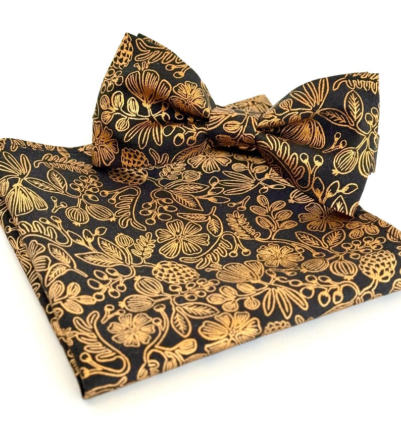 Puede incluir: Una pajarita y un pa&ntilde;uelo de bolsillo negros con un patr&oacute;n floral dorado.