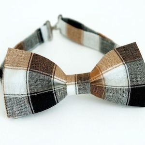 May include: A pre-tied bow tie featuring a plaid pattern in shades of brown, black, white, and gray. The fabric has a visible woven texture. The bow tie includes an adjustable fabric neck strap with a silver-toned metal sliding buckle.