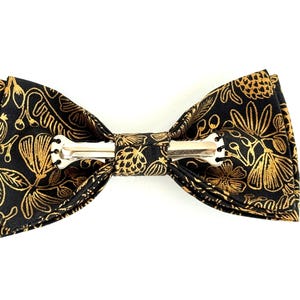 Puede incluir: Una pajarita negra con un patr&oacute;n floral dorado. La pajarita tiene un clip plateado.
