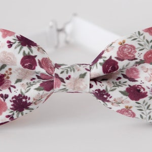 May include: A bow tie featuring a white background with a floral pattern in shades of burgundy, dusty rose, and muted green foliage. The bow tie has a central fabric knot and includes an adjustable white strap with a silver-toned metal fastener.