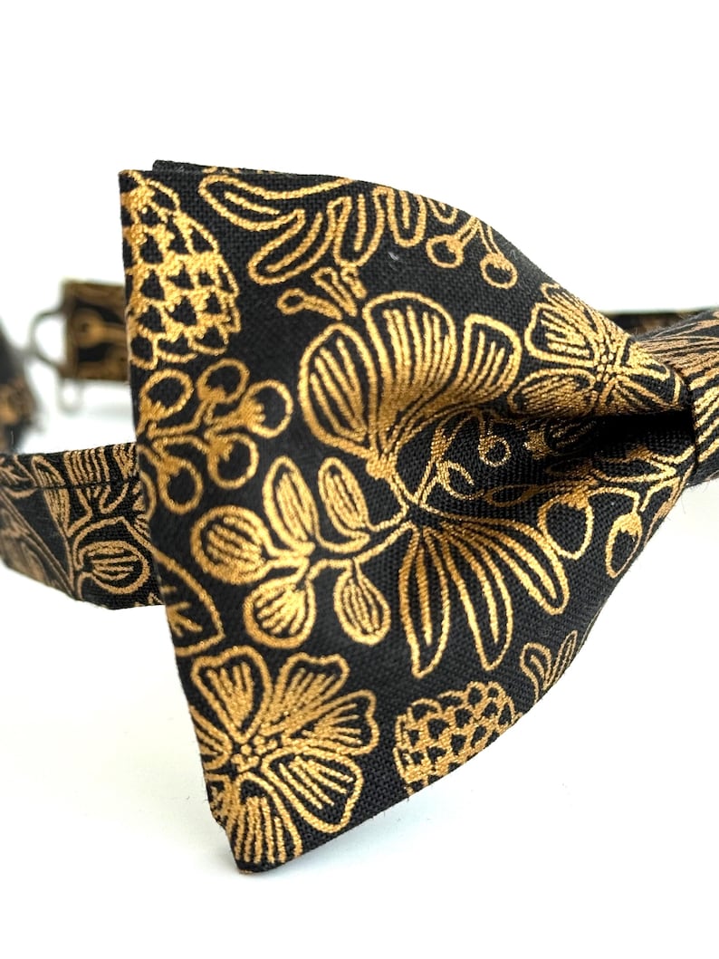Puede incluir: Una pajarita negra con un patr&oacute;n floral dorado. La pajarita est&aacute; hecha de una tela con una textura sutil.