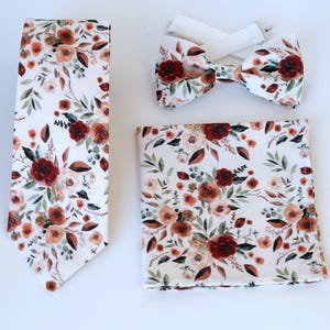 May include: A set of three matching accessories featuring a floral pattern with red, orange, and white flowers on a white background. The set includes a tie, a bow tie, and a pocket square.