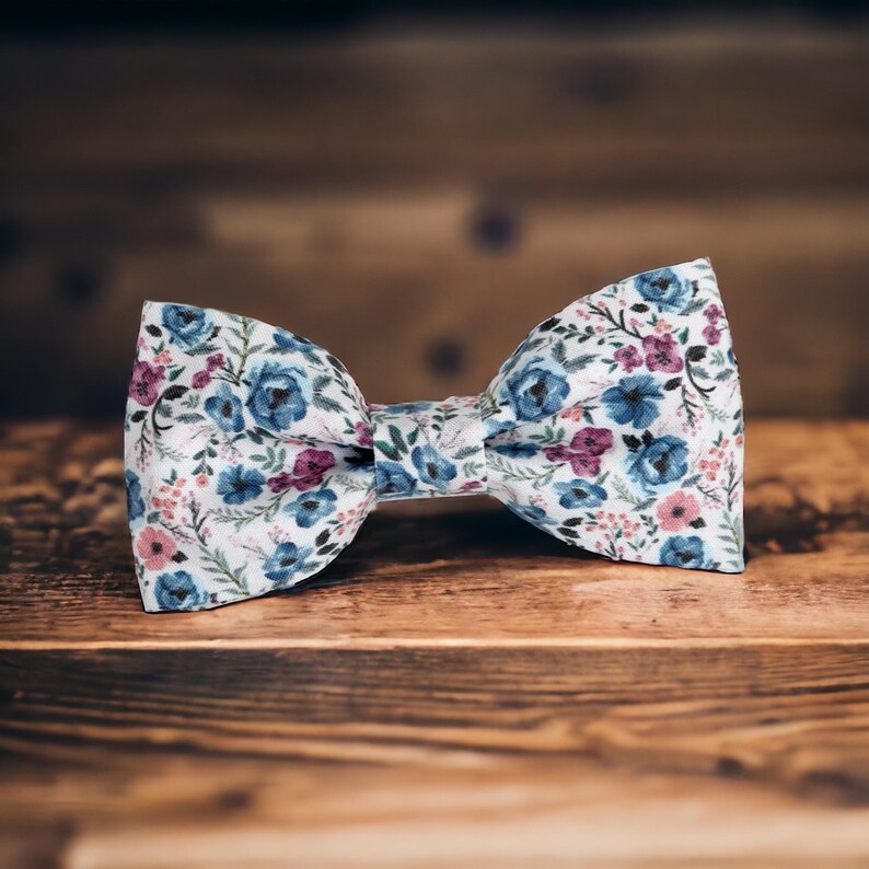 May include: A white bow tie with a floral pattern featuring blue, pink, and green flowers. The bow tie is made of fabric and has a classic bow shape.