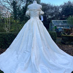 May include: A vintage ivory wedding gown with an off-the-shoulder neckline and a full skirt. The bodice features a lace-up back detail. The dress is displayed on a mannequin outdoors, with a long train and a flowing silhouette. The dress is made of a satin-like material.