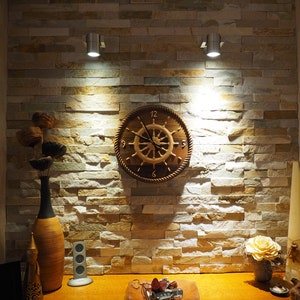 May include: A wooden ship's wheel clock hangs on a textured stone wall, illuminated by two spotlights. The clock features a light-colored wheel design with a rope-like border. A wooden table displays decorative items, including a vase, a small speaker, and a leather tray with keys.