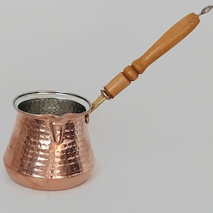Könnte beinhalten: Eine türkische Kaffeekanne aus gehämmertem Kupfer mit einem Holzgriff. Die Kanne hat ein silberfarbenes Innenleben.
