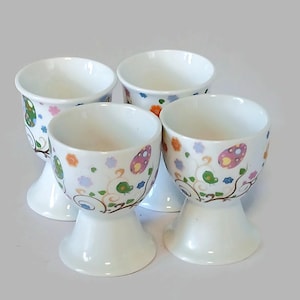 Op de afbeelding: Vier witte keramische eierdopjes met een voetstuk. Elke beker is versierd met kleurrijke bloemen- en paddenstoelontwerpen. De eierdopjes zijn perfect voor het serveren van eieren bij het ontbijt of de brunch.