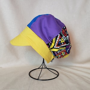 May include: A yellow, purple, and blue patterned cap with a geometric design. The cap has a wide brim and is displayed on a black stand.