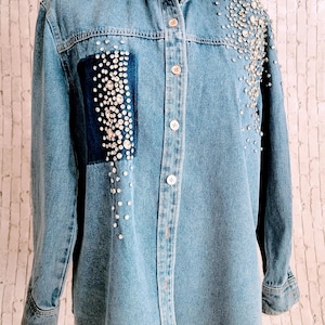 May include: Light blue denim shirt embellished with sparkling rhinestones. The shirt features a dark blue patch on the front, also adorned with rhinestones. The long-sleeved shirt has a classic collar and button-down closure.