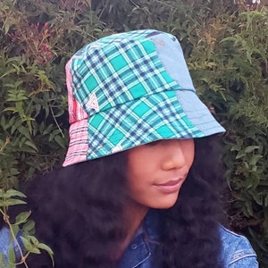 Op de afbeelding: Een persoon draagt ​​een blauwe, groene en roze geruite bucket hat met een driehoekig logo. De hoed is gemaakt van patchwork stof.