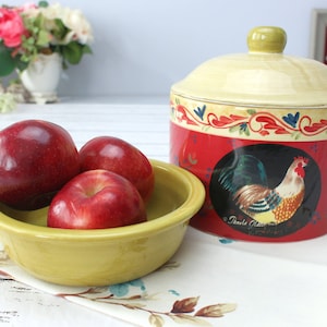 Op de afbeelding: Een keramische pot met een haanontwerp en een geel deksel. De pot is rood met een zwarte en gele haan op de voorkant. De pot staat op een wit oppervlak met een kom met rode appels.