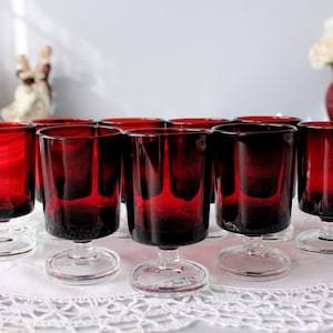 Puede incluir: Conjunto de diez vasos de chupito de vidrio rojo con tallos y bases de vidrio transparente. Los vasos están dispuestos en dos filas de cinco sobre un mantel de encaje blanco.