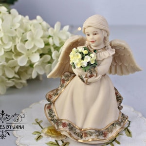 Puede incluir: Una figurita de porcelana de un ángel con alas, que lleva un vestido blanco con ribetes dorados y sostiene un ramo de flores amarillas. El ángel tiene una expresión dulce y lleva un collar de piedras preciosas verdes.