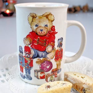 May include: A white ceramic mug with a teddy bear design in a red jacket and toy soldiers. The mug is on a white plate with two biscotti biscuits. The mug has a large handle and is decorated with a festive holiday theme.