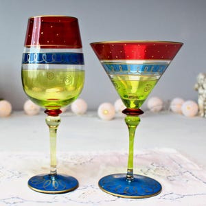 Könnte beinhalten: Zwei dekorative Weingläser. Eines ist ein Weinglas mit Stiel, das andere ein Martiniglas. Beide haben einen roten Deckel mit goldenen Punkten, einen blauen Streifen und eine gelbe Schale. Die Stiele sind gelb mit einem roten Akzent, und die Böden sind blau mit goldenen Details.