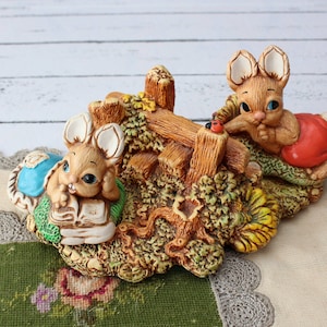 May include: Two ceramic bunny figurines sitting on a brown, leafy base. One bunny is reading a book and the other is holding a red flower.