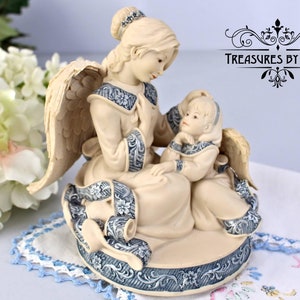 Puede incluir: Una figura de ángel color crema con un ángel niño sentado en su regazo. Ambas figuras visten túnicas a juego con adornos azules y plateados. La base y el pergamino presentan diseños similares en azul y plata. El texto "TREASURES BY JANA" es visible.