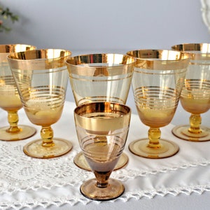 Peut inclure: Ensemble de six verres à vin en verre ambré avec une bordure dorée et un petit verre à liqueur en verre ambré avec une bordure dorée. Les verres sont sur une nappe en dentelle blanche.