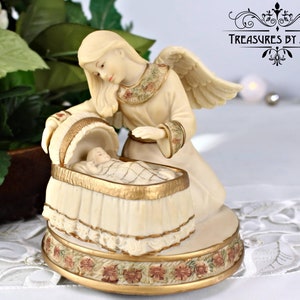 Puede incluir: Una figurita de porcelana blanca de un ángel con alas, arrodillado junto a un bebé en una cuna. El ángel lleva un vestido blanco con ribetes dorados y la cuna está decorada con detalles dorados y blancos. La figurita está sobre una base con un diseño floral.
