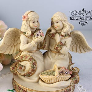 Puede incluir: Una figura de ángel color crema con dos ángeles con alas, sosteniendo ramos y una cesta de flores. Los ángeles visten vestidos largos con detalles florales. La base está decorada con motivos florales y ribetes dorados. El texto "Treasures by Jana" es visible.