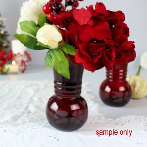 May include: Two small, dark red glass vases with artificial flowers. One vase has white and red flowers, the other has red flowers. The flowers include red hydrangeas, white peonies, and red berries. The vases are on a white lace tablecloth.