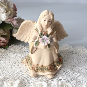 Puede incluir: Una figurita de porcelana de un ángel con alas, sosteniendo un ramo de flores moradas y blancas. El ángel lleva un vestido color crema con detalles dorados y está sentado sobre un encaje de ganchillo blanco.