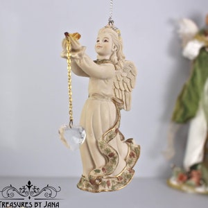 Puede incluir: Una figurita de ángel de porcelana blanca con detalles dorados y un corazón de cristal colgando de una cadena dorada. El ángel sostiene un pequeño pájaro en su mano y tiene un diseño floral en su vestido.