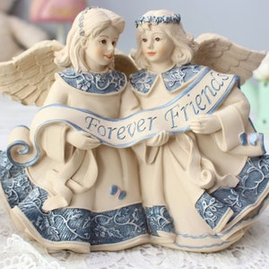 Puede incluir: Figura de color crema de dos ángeles sosteniendo una pancarta que dice "Forever Friends" en escritura azul. Los ángeles tienen alas, halos y visten túnicas azules y crema con detalles florales. Una pieza decorativa.