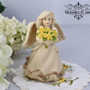 Puede incluir: Una figura de ángel color crema que sostiene un ramo de flores amarillas. El ángel tiene alas emplumadas y un vestido largo con un ribete dorado decorativo. La figura está sobre un tapete blanco con bordados florales amarillos. El texto "Treasures by Jana" es visible.