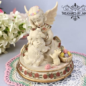 Puede incluir: Una figurita de porcelana de un ángel con alas, una corona de flores y un vestido blanco, sosteniendo un perro blanco. El ángel está sentado sobre una base con una cesta de lana y un gato. La base está decorada con flores y detalles dorados. "Treasures by Jana" está escrito en la base.
