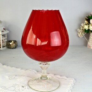 May include: A large, vibrant red brandy glass with a clear, decorative stem and base. The glass has a rounded bowl and a scalloped rim. The glass sits on a white, embroidered doily, with a soft background.