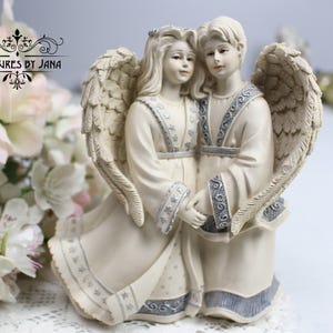 Puede incluir: Una figura de ángel color crema que representa dos figuras tomadas de la mano. Los ángeles tienen alas emplumadas y visten túnicas con adornos azules y plateados. El texto "Treasures by Jana" es visible en la esquina superior izquierda.