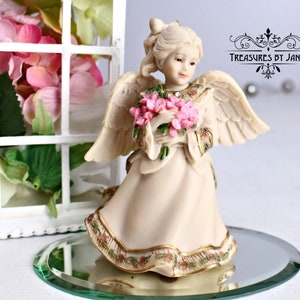 Puede incluir: Una figurita de porcelana de un ángel sosteniendo un ramo de flores rosas. El ángel lleva un vestido blanco con ribetes dorados y una corona de flores en la cabeza. La figurita está sentada sobre una base redonda de espejo.