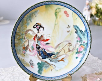 May include: A decorative porcelain plate with a gold rim and a blue and white border. The plate features a hand-painted image of a woman in a pink and blue dress with a fan in her hand. The woman is surrounded by flowers and foliage, and the background is a yellow and green landscape. The plate is on a stand.