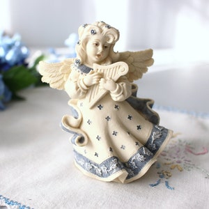 Puede incluir: Figura de ángel color crema tocando un arpa. El ángel tiene alas detalladas y viste un vestido con detalles azules y pequeños diseños en forma de cruz. La figura está sobre una superficie blanca con detalles florales azules.