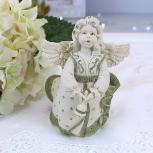 Puede incluir: Una figurita de ángel de porcelana blanca con alas verdes y blancas, un vestido verde y blanco y una campana en la mano. El ángel tiene una dulce expresión en su rostro y está mirando hacia arriba.